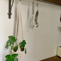 Indoor cream cotton hanging plant holder with three long crocheted straps, finished with a loop to hang up. Image shows planter hanging in a kitchen, from a hanging rail, with a pot and plant inside. Made with recycled cotton cord.