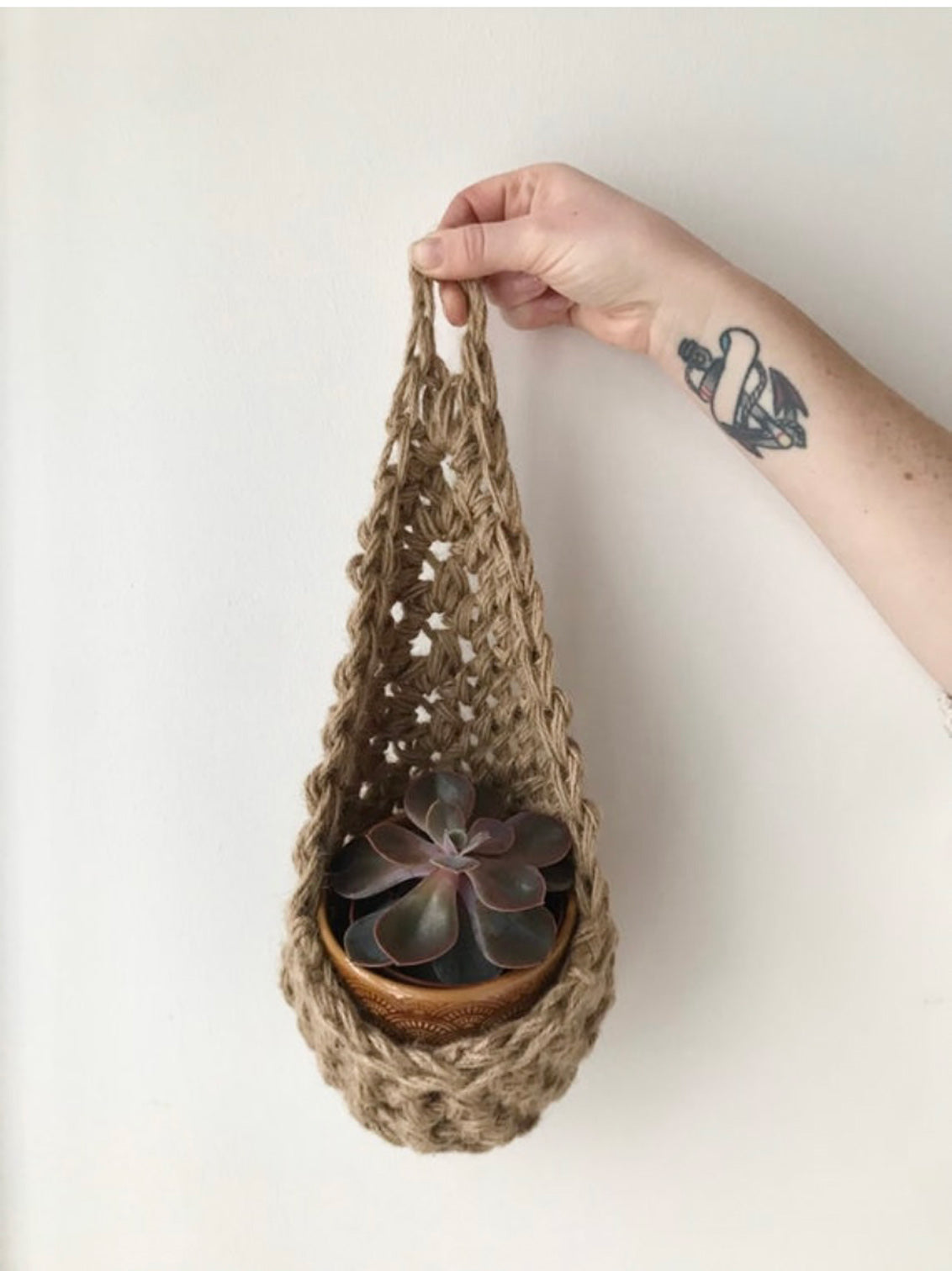 Photo showing a single handmade natural jute wall planter, being held front on by makers hand. Showing it's teardrop shape with ceramic pot and plant inside it.