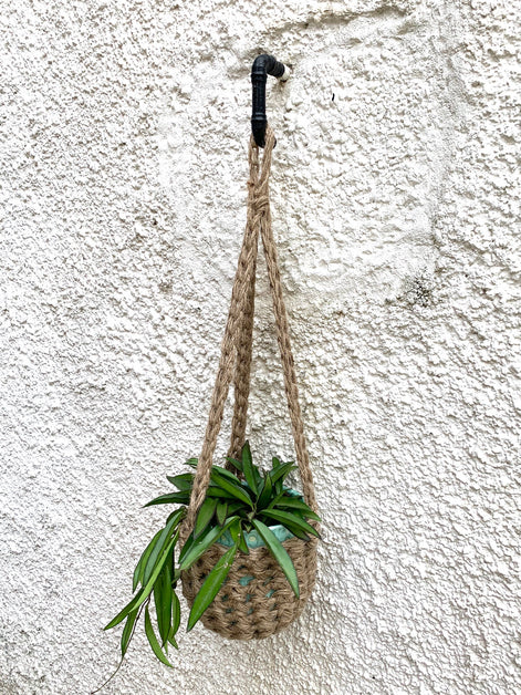 Natural Jute hanging plant holder with three long crocheted straps and pot shaped basket at the bottom of these straps designed for pot to be placed securely, finished with a loop to hang up. Image shows planter outside, suspended from a small hook.