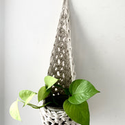 Image of single white cotton crochet hanging wall planter against a wall of a bedroom. The photo shows the planter with a pot and plant inside it, in order to demonstate tierdrop shape of design. Hanging by a small nail in the wall.