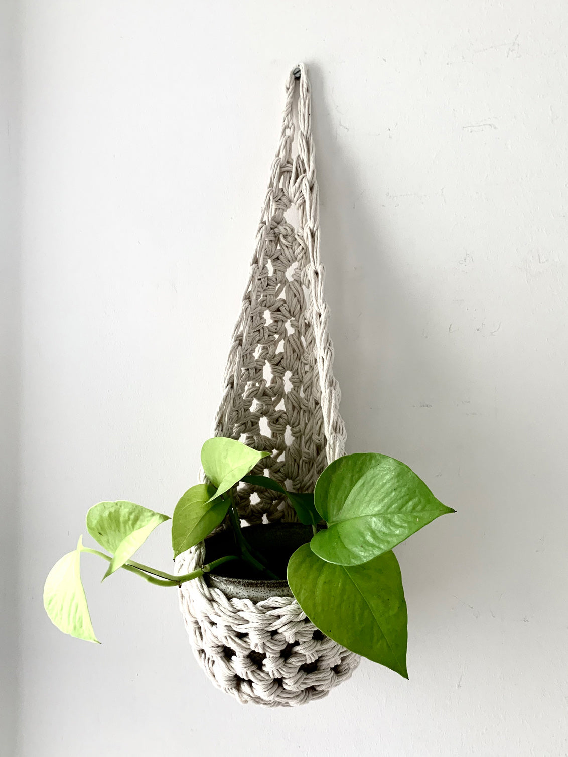 Image of single white cotton crochet hanging wall planter against a wall of a bedroom. The photo shows the planter with a pot and plant inside it, in order to demonstate tierdrop shape of design. Hanging by a small nail in the wall.