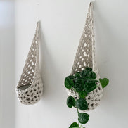 Image of two white cotton crochet hanging wall planter against a wall of a bedroom. The photo shows the planter with a pot and plant inside it, in order to demonstate tierdrop shape of design. Hanging by a small nail in the wall.
