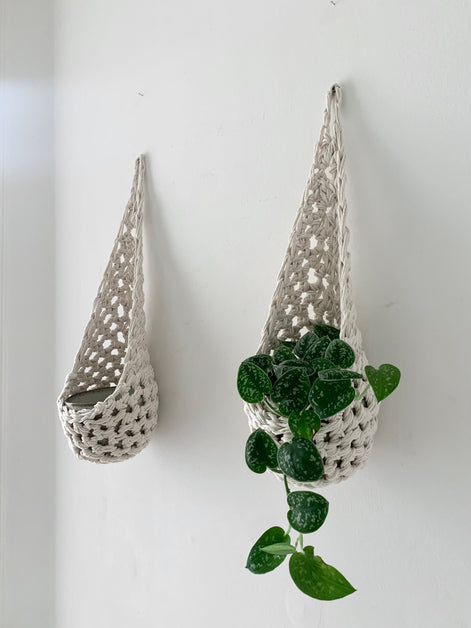 Image of two white cotton crochet hanging wall planter against a wall of a bedroom. The photo shows the planter with a pot and plant inside it, in order to demonstate tierdrop shape of design. Hanging by a small nail in the wall.