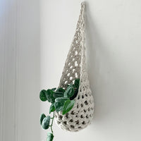 Image of single white cotton crochet hanging wall planter against a wall of a bedroom. The photo shows the planter with a pot and plant inside it, in order to demonstate tierdrop shape of design. Hanging by a small nail in the wall.