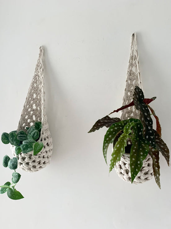 Image of two white cotton crochet hanging wall planter against a wall of a bedroom. The photo shows the planter with a pot and plant inside it, in order to demonstate tierdrop shape of design. Hanging by a small nail in the wall.