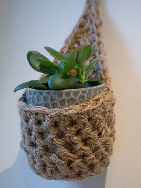  Photo showing a single handmade crochet jute hanging wall planter, against a white wall. The planter is a tierdrop shape and is holding a pot and plant